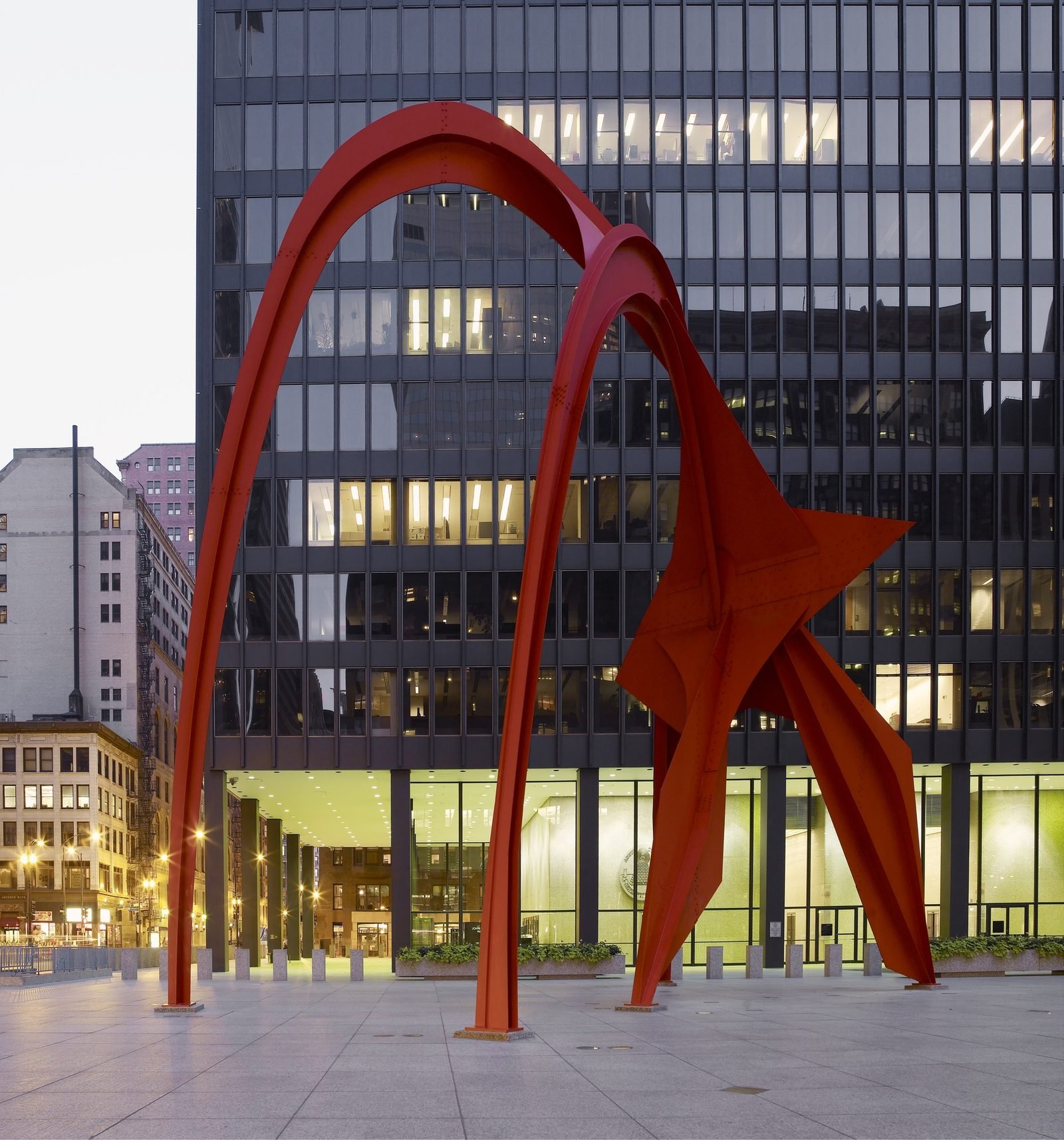 A dark steel and glass office tower above a city plaza, with a red steel sculpture at its foot